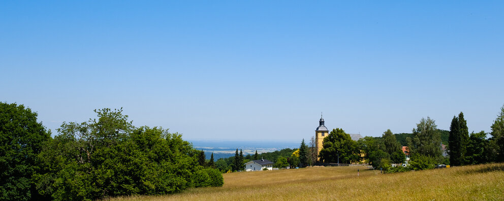 Ausblick über Neunkirchen, die Frankfurter Skyline bis zum Taunus. Im Vordergrund sind eine Wiese und hinter ein paar Bäumen ein Kirchturm zu sehen.